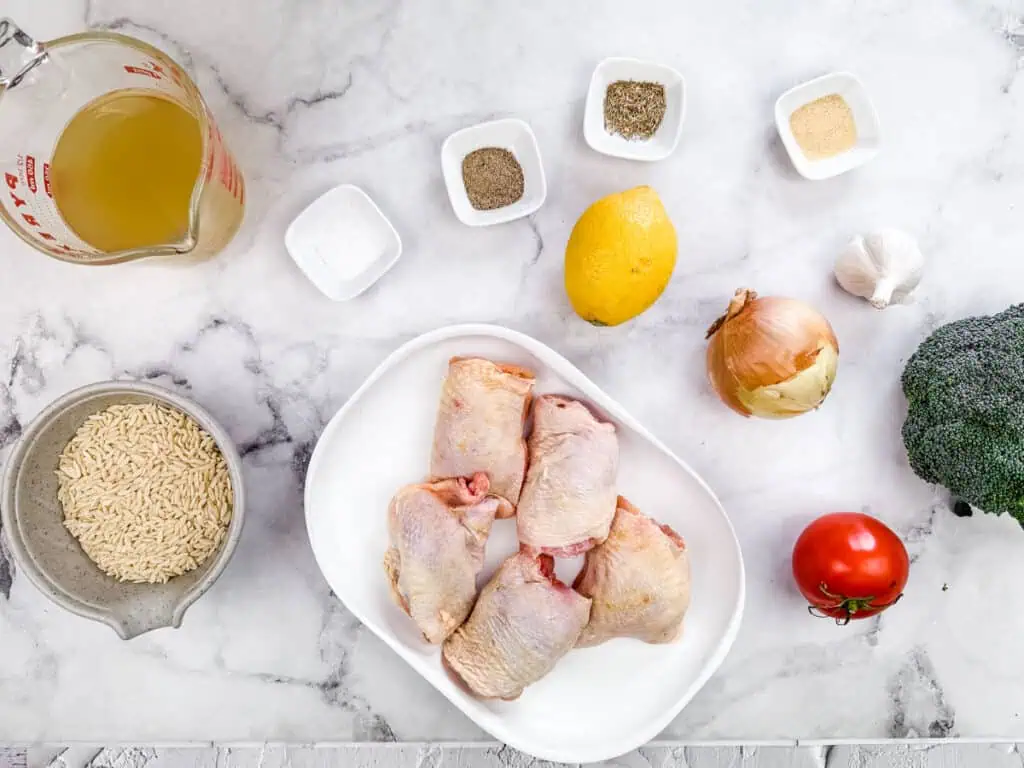 Flat lay of fresh ingredients for a Lemon Chicken Orzo Skillet with Broccoli, including raw chicken thighs, orzo pasta, chicken broth, lemon, broccoli, tomato, onion, garlic, and seasonings on a marble countertop.