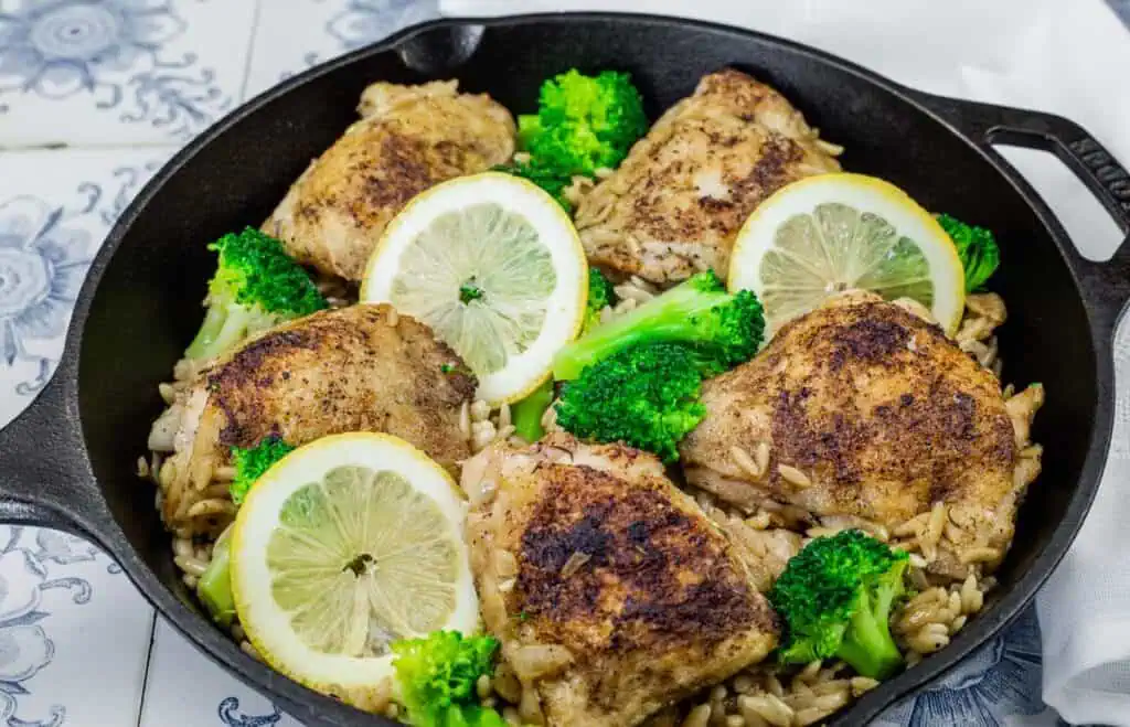 Golden-browned lemon chicken thighs served over orzo pasta with tender broccoli florets, garnished with fresh lemon slices in a cast-iron skillet.