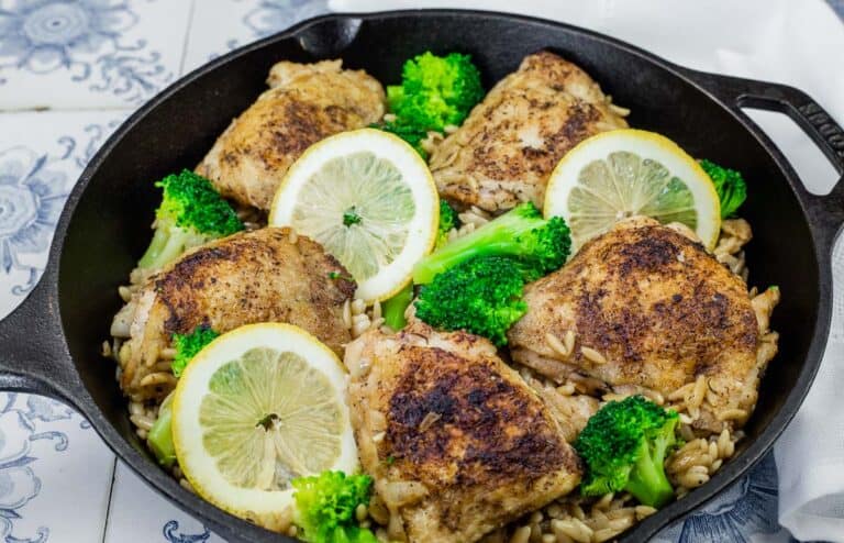 Golden-browned lemon chicken thighs served over orzo pasta with tender broccoli florets, garnished with fresh lemon slices in a cast-iron skillet.