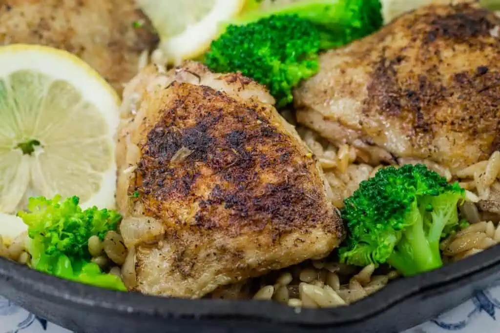 Close-up of crispy, golden-browned lemon chicken thigh atop a bed of orzo pasta, garnished with fresh broccoli florets and lemon slices in a cast-iron skillet.