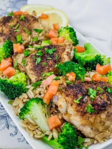 A Lemon Chicken Orzo Skillet with Broccoli dish featuring golden-brown seasoned chicken thighs on a bed of orzo pasta, garnished with fresh broccoli florets, diced tomatoes, chopped parsley, and lemon slices in the background.