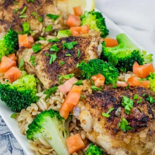 A Lemon Chicken Orzo Skillet with Broccoli dish featuring golden-brown seasoned chicken thighs on a bed of orzo pasta, garnished with fresh broccoli florets, diced tomatoes, chopped parsley, and lemon slices in the background.