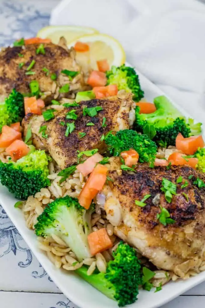 A Lemon Chicken Orzo Skillet with Broccoli dish featuring golden-brown seasoned chicken thighs on a bed of orzo pasta, garnished with fresh broccoli florets, diced tomatoes, chopped parsley, and lemon slices in the background.
