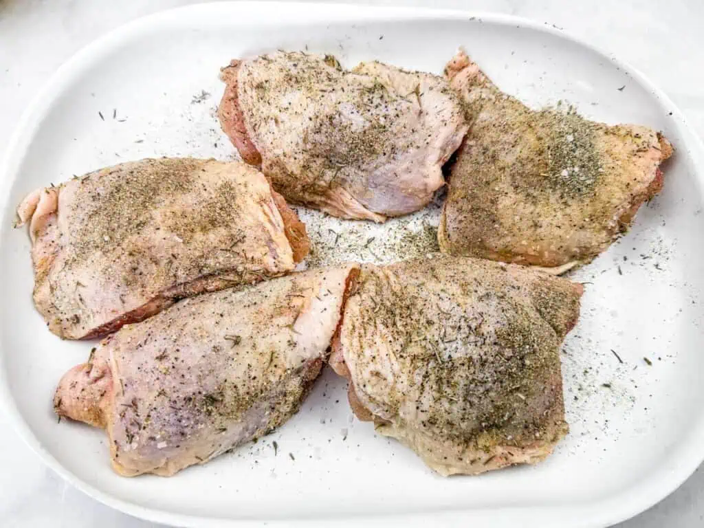 Raw chicken thighs seasoned with a blend of herbs and spices, arranged on a white plate, ready for cooking.
