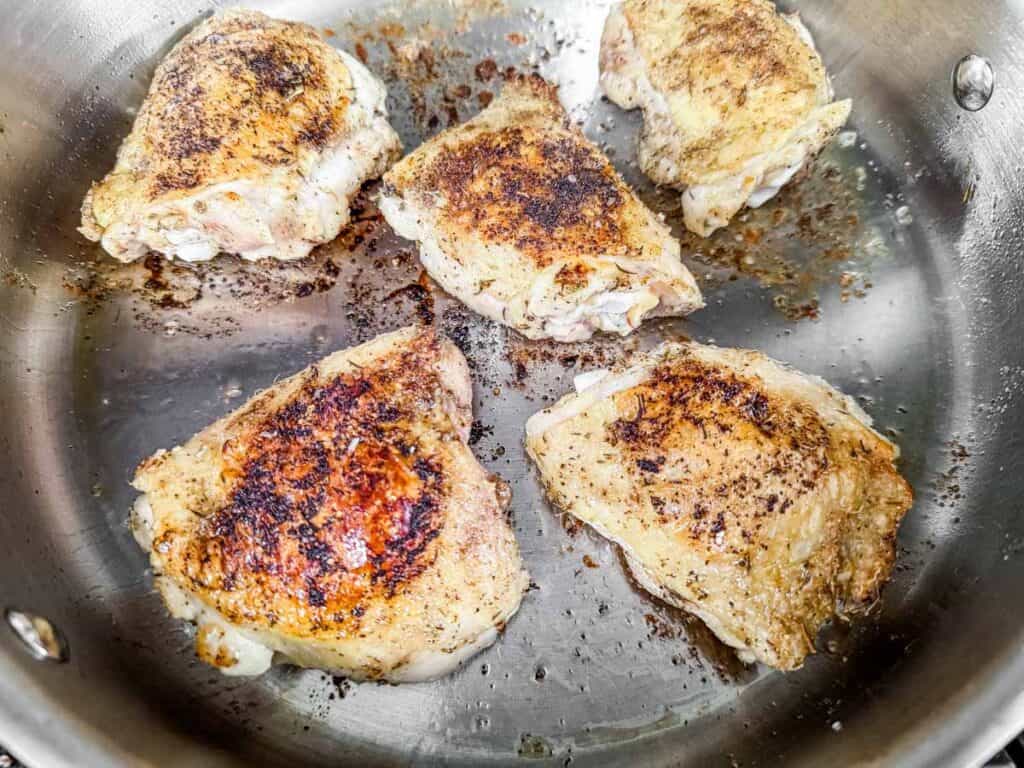 Chicken thighs being seared in a stainless steel pan, developing a golden-brown, crispy skin with flavorful seasonings.