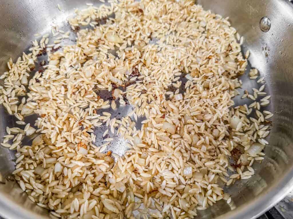 Orzo pasta and saut&eacute;ed onions toasting in a stainless steel pan, absorbing rich flavors from the browned bits left behind.