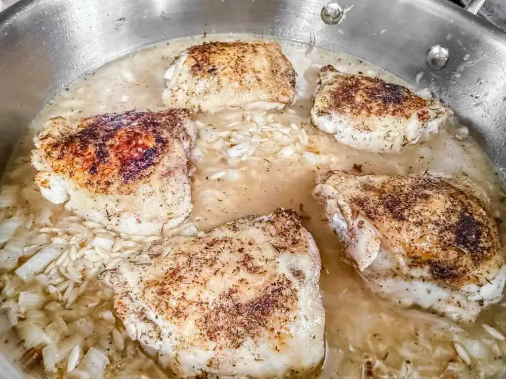 Seared chicken thighs simmering in a flavorful broth with orzo pasta and onions in a stainless steel skillet.