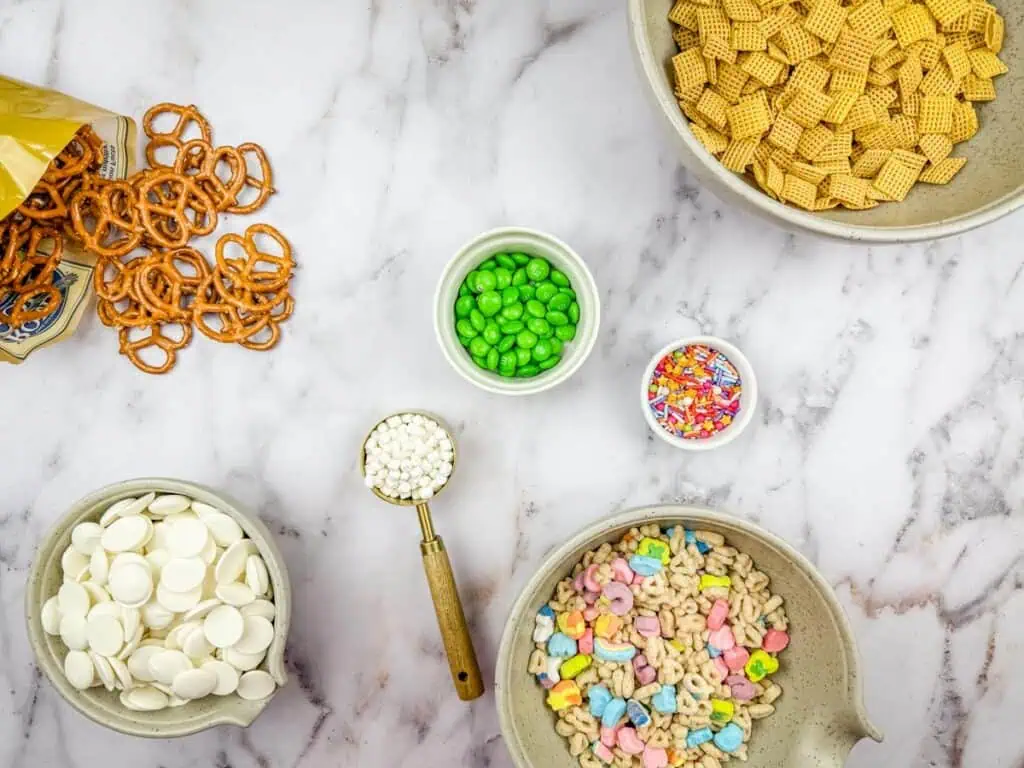 Ingredients for a snack mix are arranged on a marble surface, including pretzels, cereal, chocolate wafers, green candy, mini marshmallows, sprinkles, and cereal with marshmallow bits.