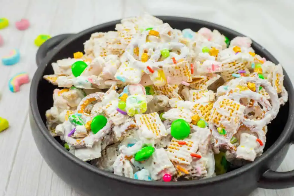 A black bowl filled with a snack mix, including cereal squares, pretzels, colorful candy pieces, and coated in a white coating.