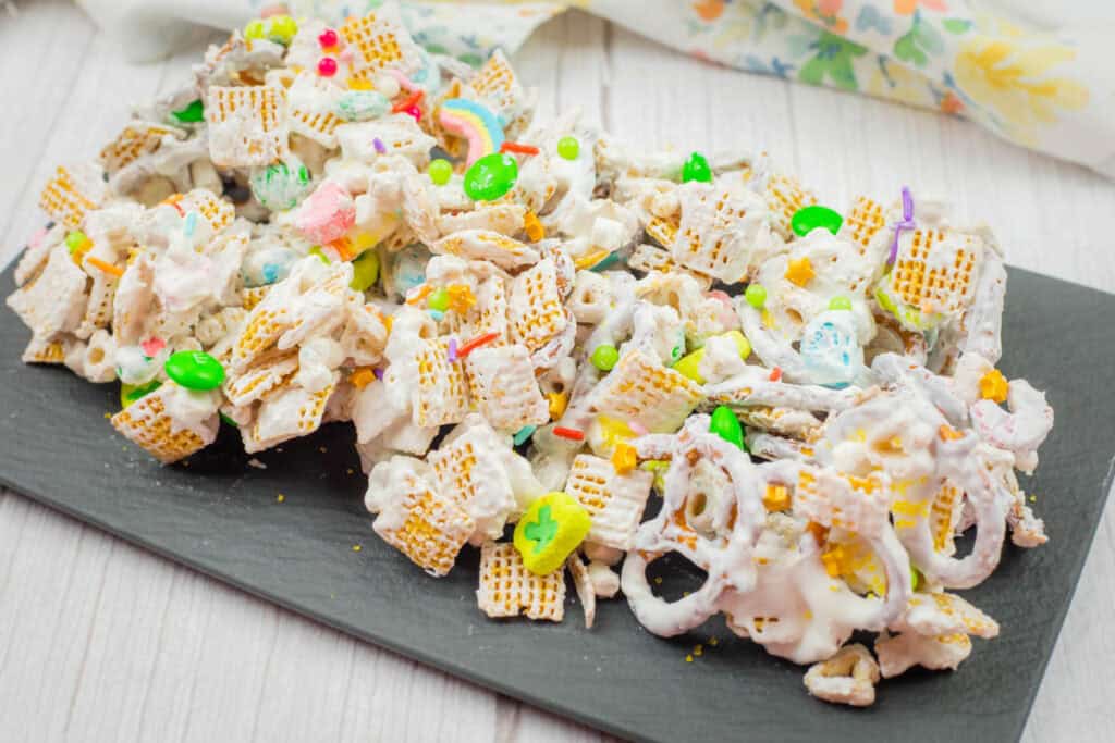A platter of white chocolate-coated snack mix with cereal, pretzels, and colorful candy on a slate board, set atop a white wooden surface.