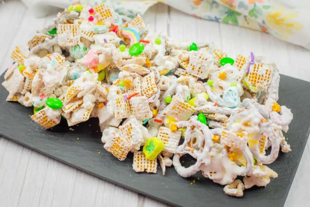 A platter of white chocolate-coated snack mix with cereal, pretzels, and colorful candy on a slate board, set atop a white wooden surface.