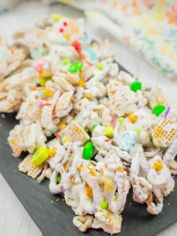Colorful snack mix with cereal pieces, pretzels, and candies coated in white frosting on a dark serving tray.