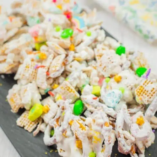 Colorful snack mix with cereal pieces, pretzels, and candies coated in white frosting on a dark serving tray.