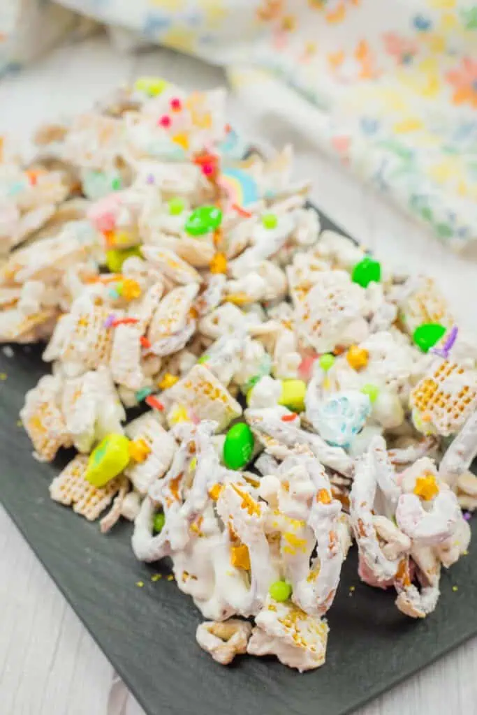 Colorful snack mix with cereal pieces, pretzels, and candies coated in white frosting on a dark serving tray.
