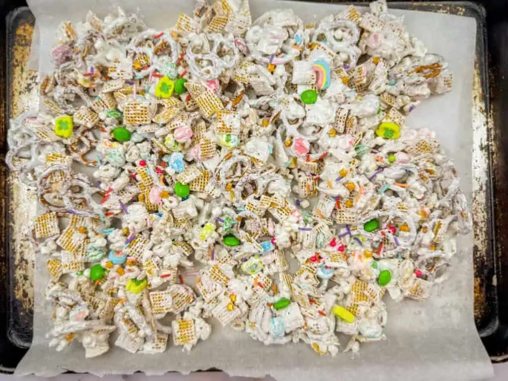 A tray of snack mix with pretzels, cereal, and colorful candies, coated in white confectionery on parchment paper.