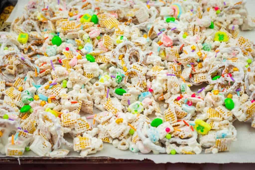 A colorful snack mix with cereal, candy pieces, and pretzels covered in white chocolate and sprinkles on a lined baking sheet.