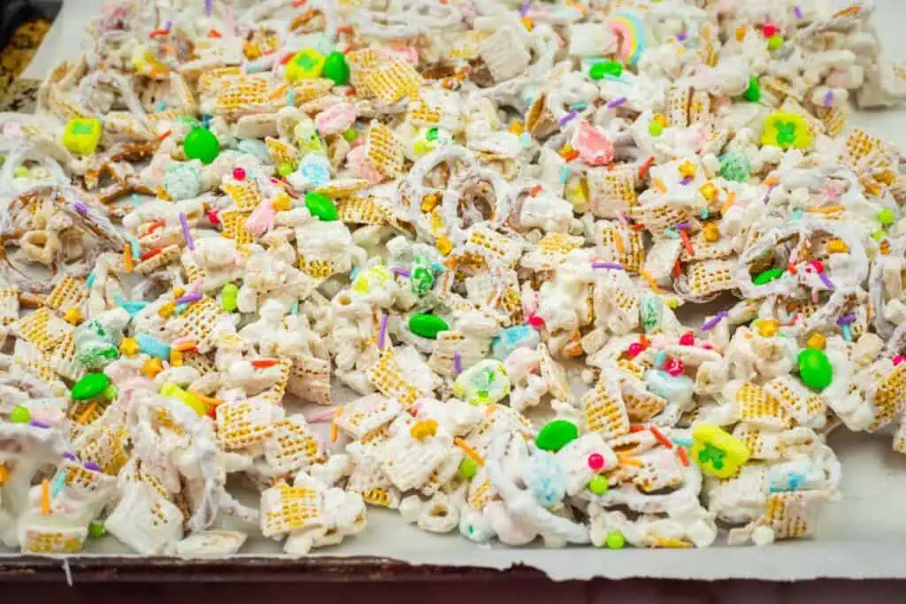 A colorful snack mix with cereal, candy pieces, and pretzels covered in white chocolate and sprinkles on a lined baking sheet.