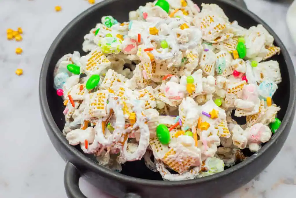 A black bowl filled with a mix of cereal, pretzels, colorful candies, and marshmallows, coated in a white, sweet glaze.