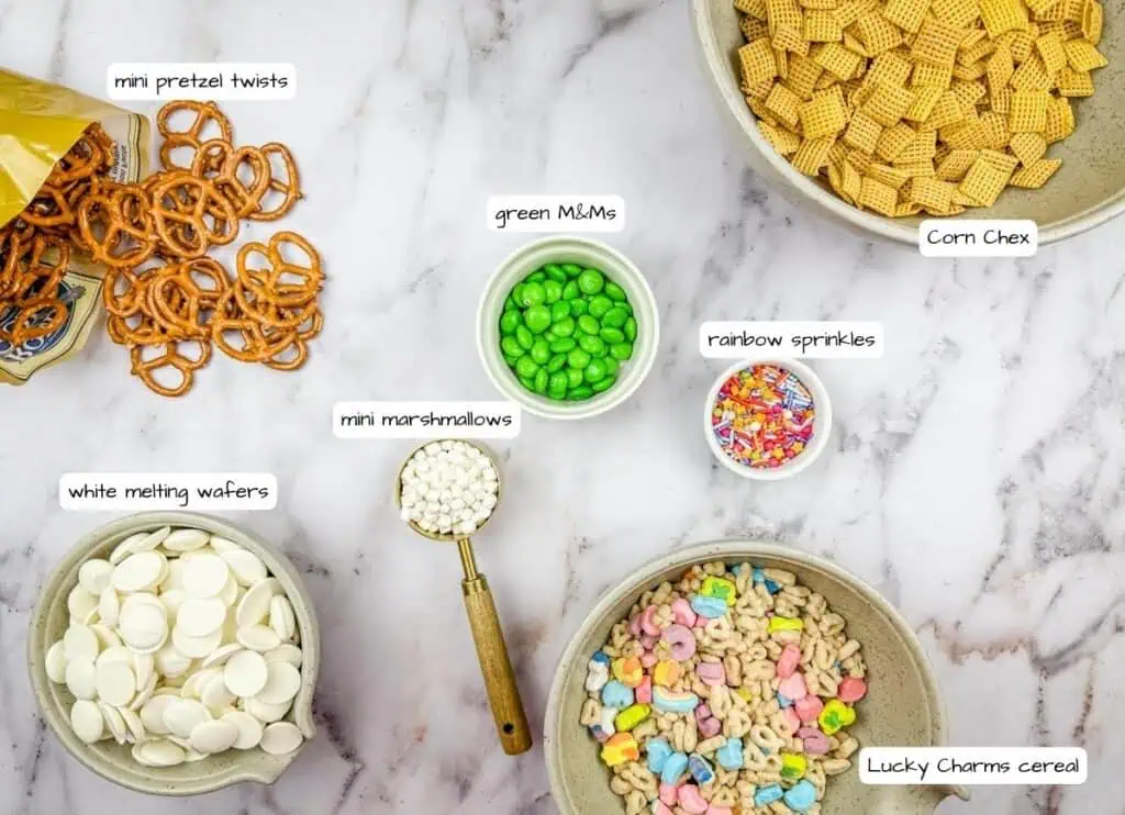 Various snack ingredients are arranged on a table, including mini pretzel twists, Corn Chex, Lucky Charms cereal, white melting wafers, mini marshmallows, green M&Ms, and rainbow sprinkles.