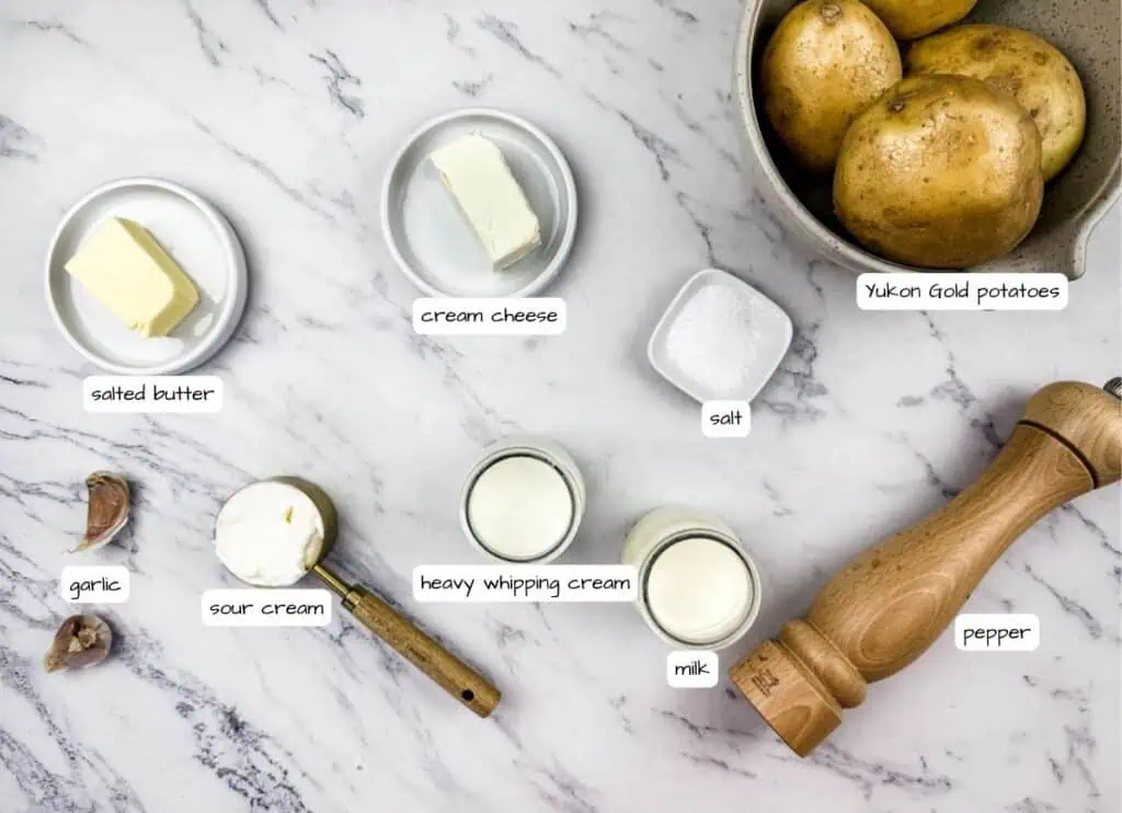 Mashed Potatoes with Cream Cheese ingredients on a marble surface, including butter, cream cheese, sour cream, garlic, milk, salt, pepper, and a bowl of potatoes with label.