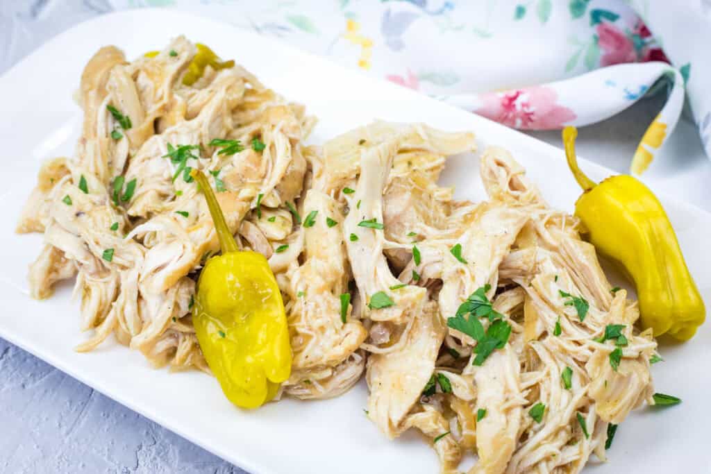 Slow Cooker Mississippi Chicken with chopped herbs and two yellow peppers on a white rectangular plate, placed on a gray surface with a floral cloth in the background.