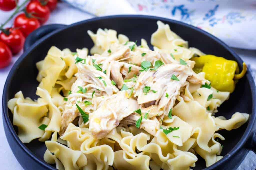 A black bowl containing egg noodles topped with Slow Cooker Mississippi Chicken and garnished with parsley, accompanied by a pickled pepper. Fresh cherry tomatoes are in the background.