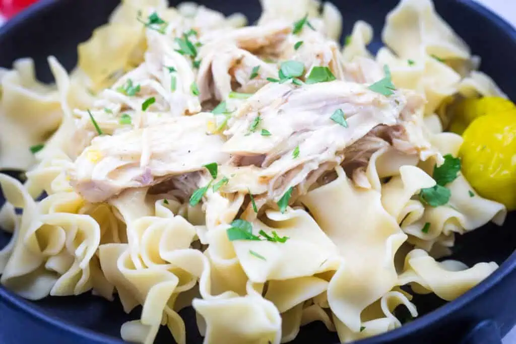 A dish of egg noodles topped with Slow Cooker Mississippi Chicken and parsley, served with a pepper.