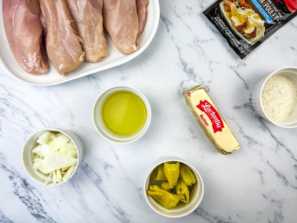 Raw chicken breasts on a plate, with small bowls of sliced onions, olive oil, chopped peppers, grated cheese, a stick of butter, and a seasoning pack on a marble surface.