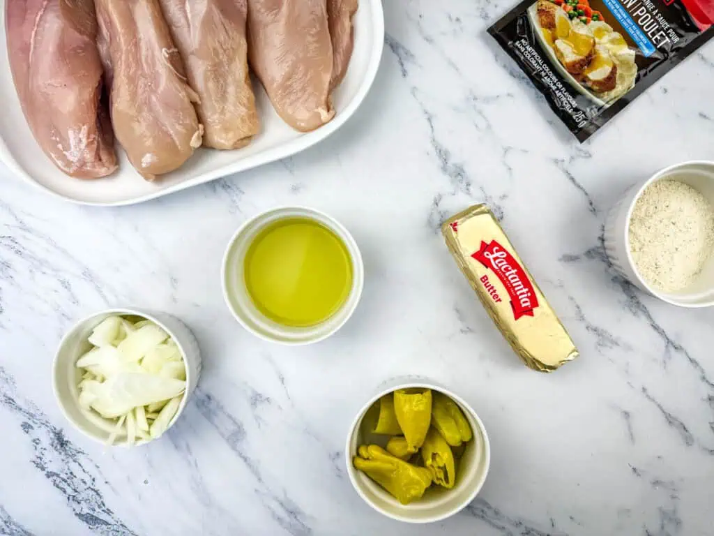 Raw chicken breasts on a plate, with small bowls of sliced onions, olive oil, chopped peppers, grated cheese, a stick of butter, and a seasoning pack on a marble surface.