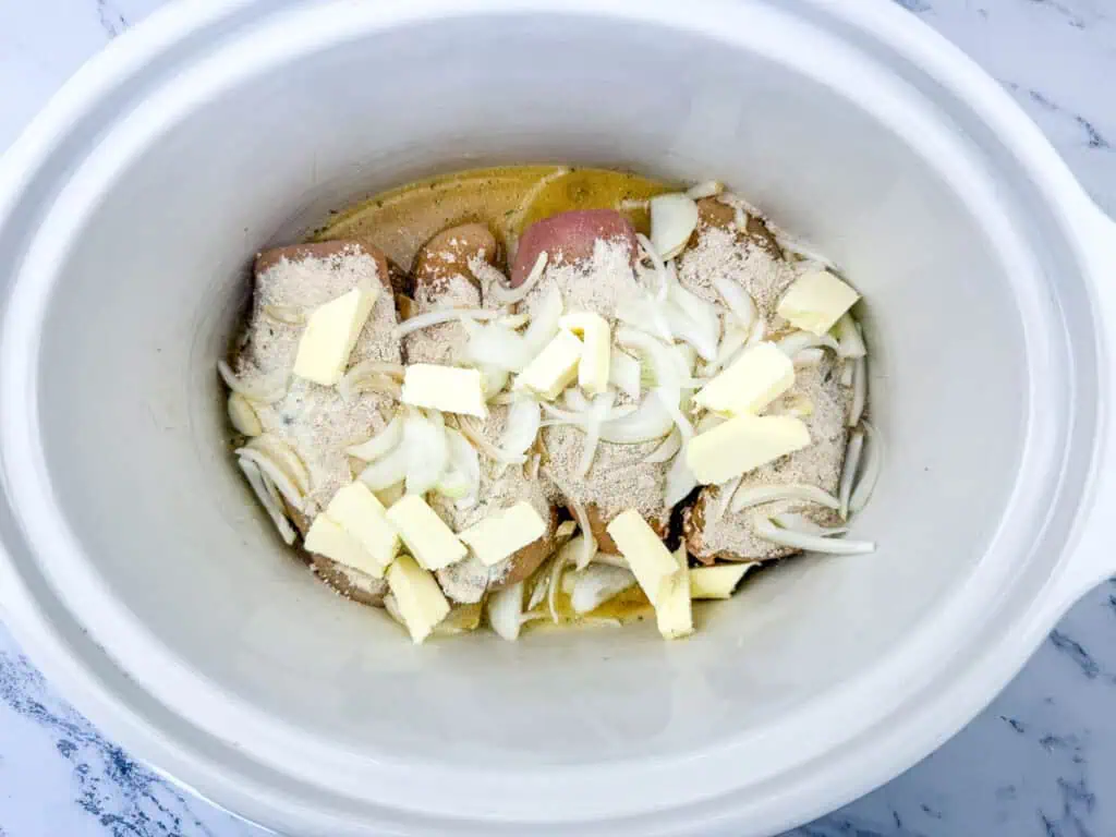 Raw chicken breasts topped with sliced onions, butter, and seasoning in a slow cooker.