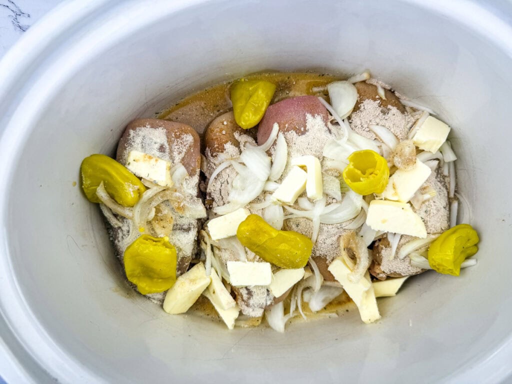 Raw chicken in a slow cooker with sliced onions, butter, pepperoncini peppers, and seasoning.
