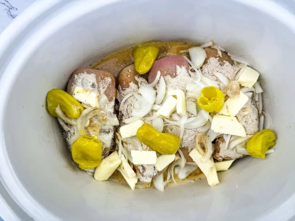 Raw chicken in a slow cooker with sliced onions, butter, pepperoncini peppers, and seasoning.