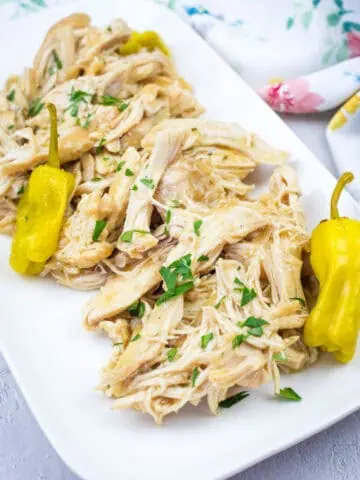 Slow Cooker Mississippi Chicken with herbs and two yellow peppers on a white rectangular plate, placed on a light surface with a floral cloth in the background.