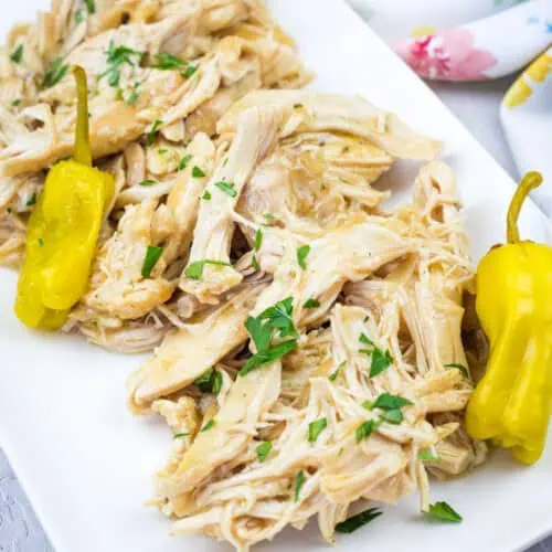 Slow Cooker Mississippi Chicken with herbs and two yellow peppers on a white rectangular plate, placed on a light surface with a floral cloth in the background.