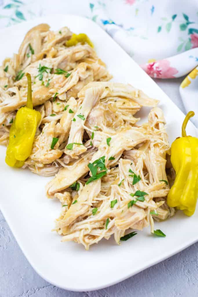 Slow Cooker Mississippi Chicken with herbs and two yellow peppers on a white rectangular plate, placed on a light surface with a floral cloth in the background.