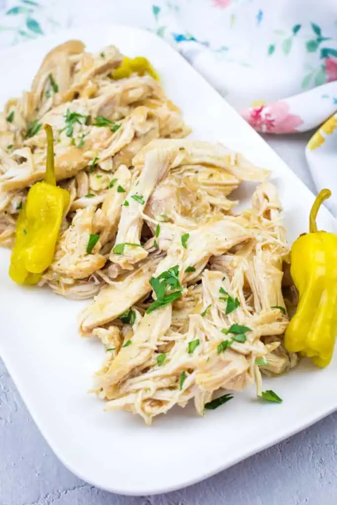 Slow Cooker Mississippi Chicken with herbs and two yellow peppers on a white rectangular plate, placed on a light surface with a floral cloth in the background.