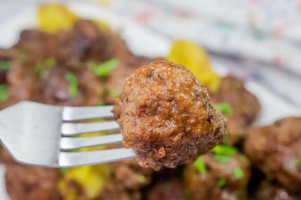 A close-up of a juicy Mississippi meatball on a fork, coated in a flavorful, rich sauce. The background features a plate of meatballs garnished with fresh parsley and accompanied by tangy pepperoncini peppers.