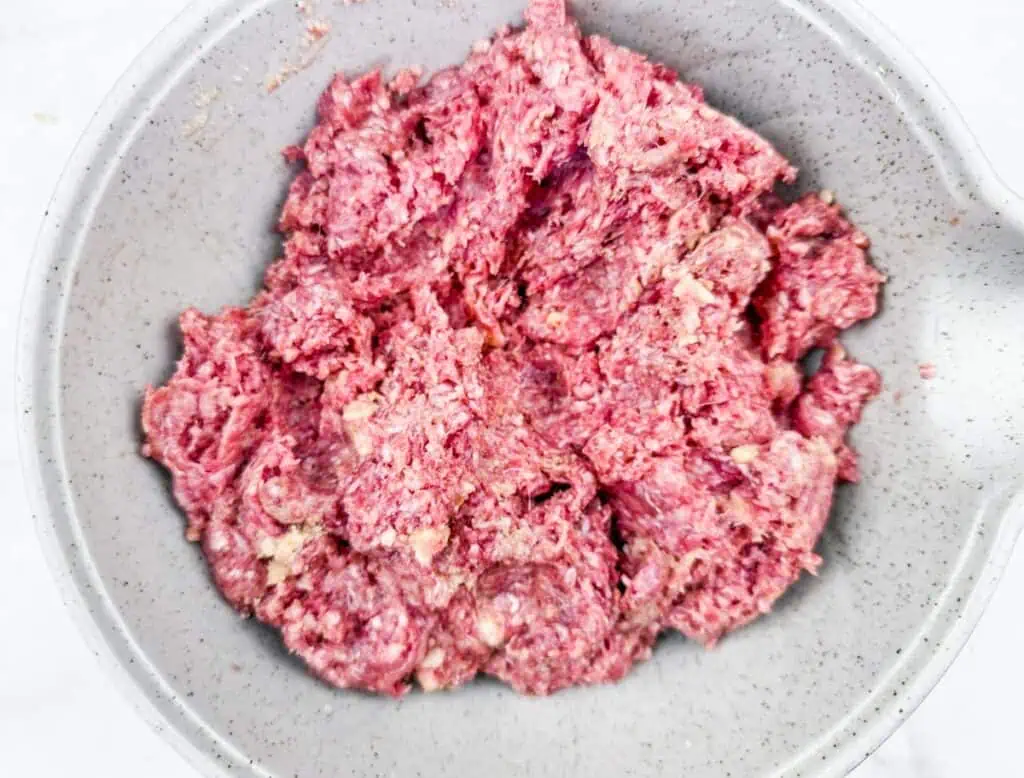 A close-up view of a mixing bowl filled with raw ground beef mixture, combined with breadcrumbs, seasonings, and other ingredients, ready to be shaped into meatballs.