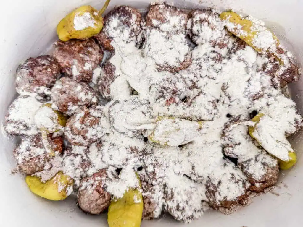 Browned meatballs and pepperoncini peppers in a slow cooker, sprinkled with a dry seasoning mix, ready for slow cooking.