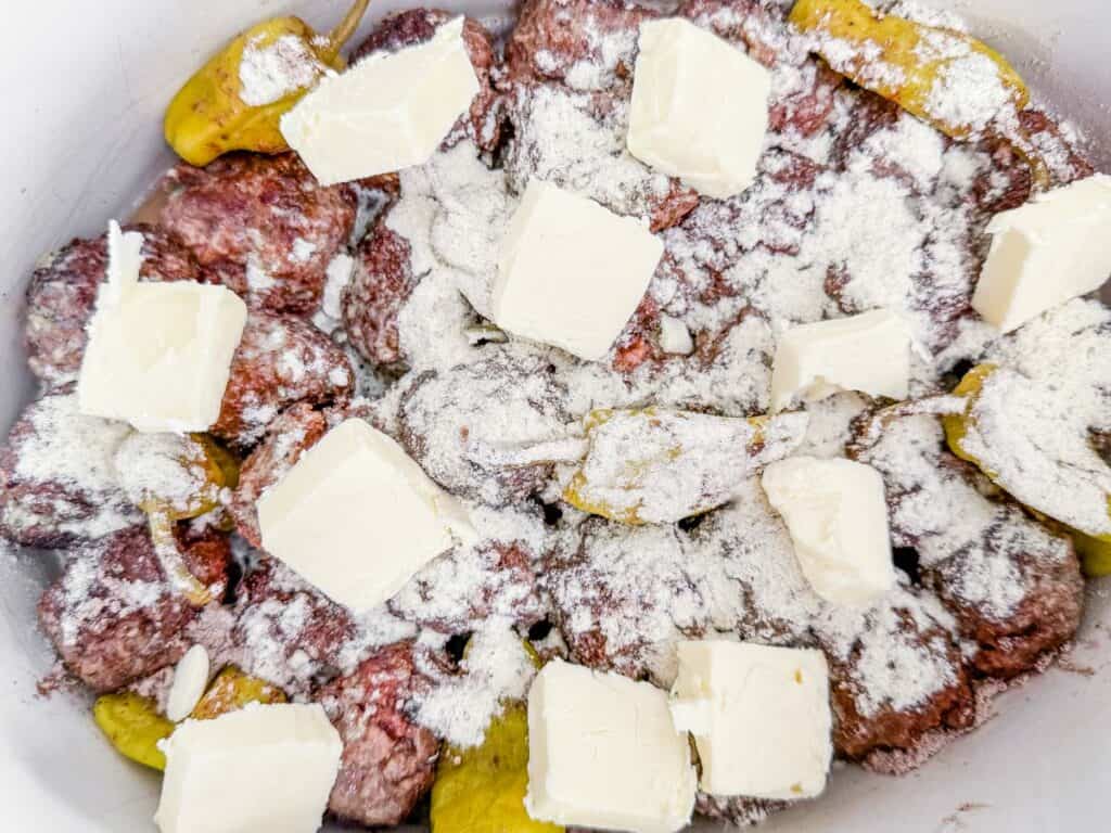 Browned meatballs and pepperoncini peppers in a slow cooker, topped with a dry seasoning mix and cubed butter, ready for slow cooking.