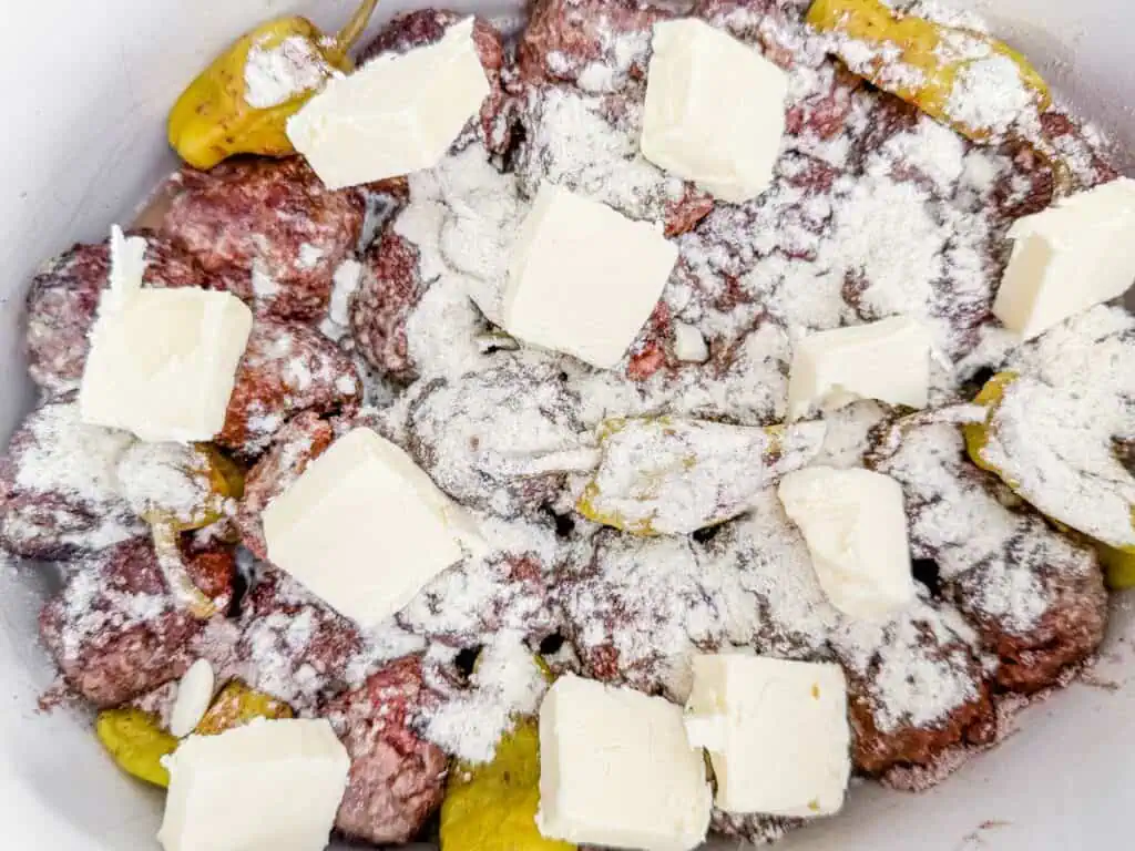 Browned meatballs and pepperoncini peppers in a slow cooker, topped with a dry seasoning mix and cubed butter, ready for slow cooking.