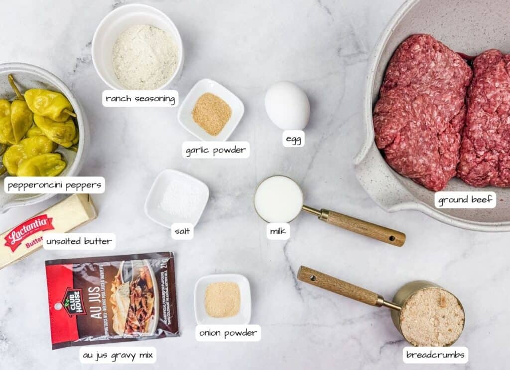 An overhead view of labeled ingredients for Mississippi meatballs, including ground beef, an egg, breadcrumbs, milk, butter, pepperoncini peppers, au jus seasoning, ranch seasoning, salt, garlic powder, and onion powder, all arranged on a white marble surface.