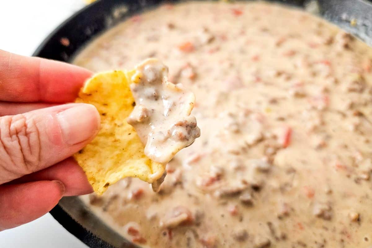 A hand holds a chip dipped in creamy Queso Blanco Rotel Dip over a pan of the same mixture.