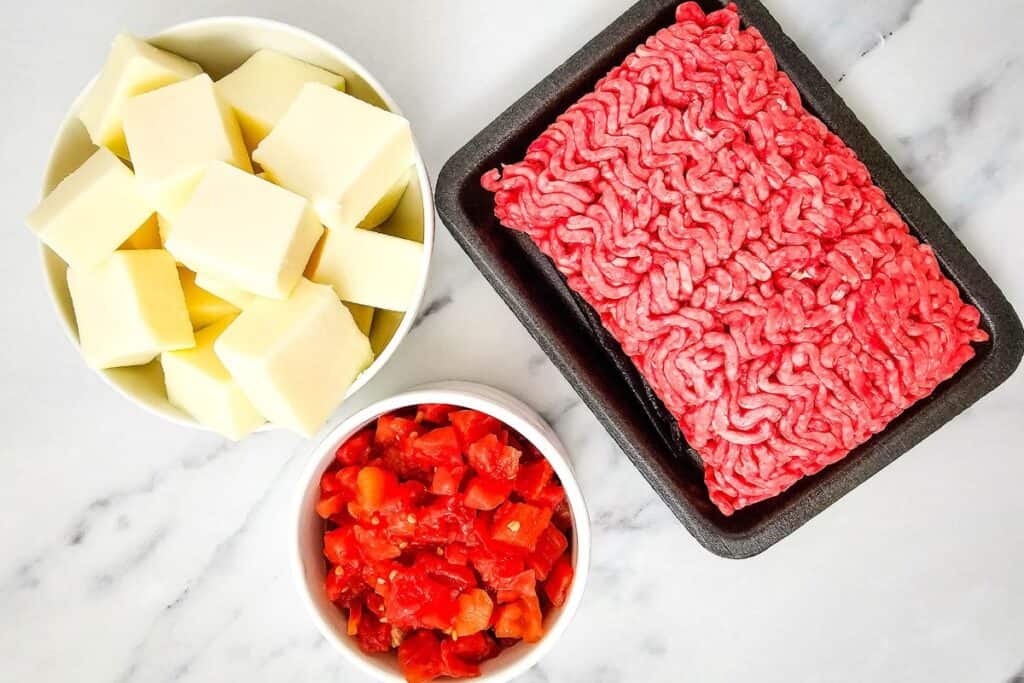 Ground beef on a tray, white bowl of cheese cubes, and a small bowl of chopped tomatoes on a marble surface.
