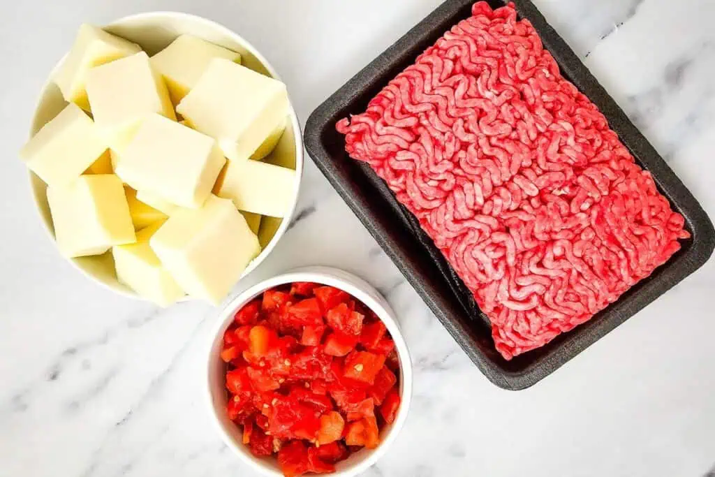 Ground beef on a tray, white bowl of cheese cubes, and a small bowl of chopped tomatoes on a marble surface.
