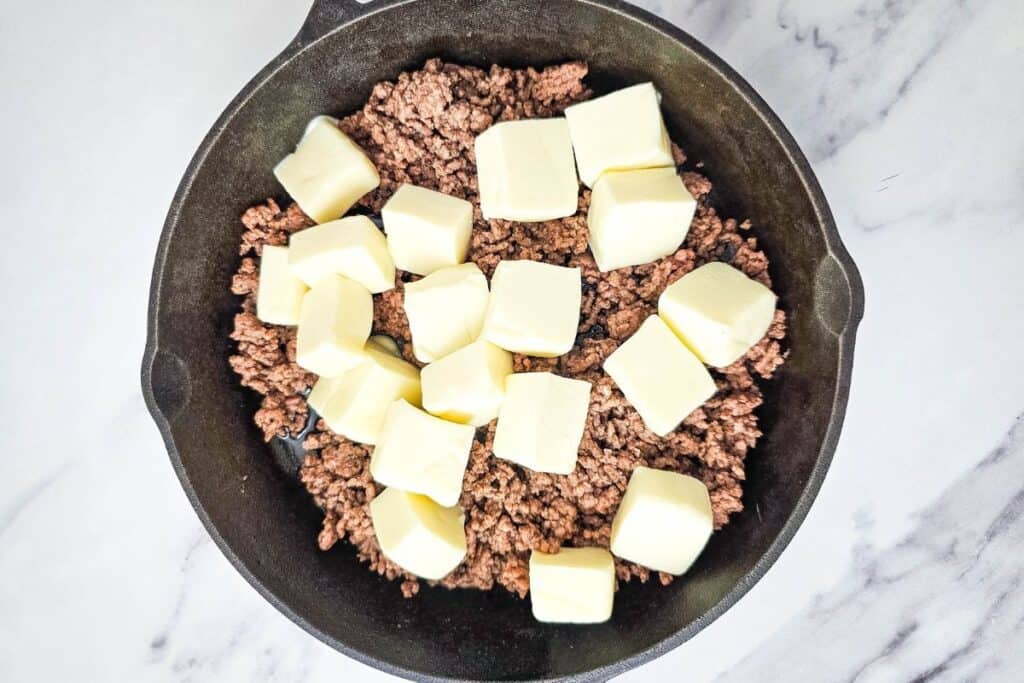 Ground meat with cubed butter in a black skillet on a marble surface.