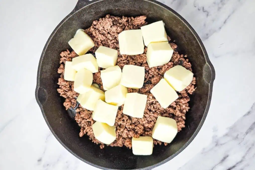 Ground meat with cubed butter in a black skillet on a marble surface.