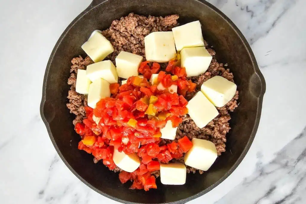 A skillet contains cooked ground beef, diced tomatoes, and several cubes of cheese on a marble surface.