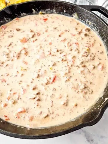 A cast iron skillet filled with creamy Queso Blanco Rotel Dip containing ground meat and diced tomatoes.