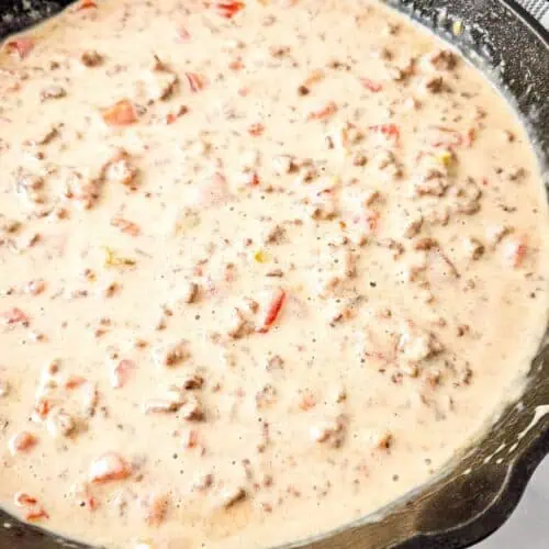 A cast iron skillet filled with creamy Queso Blanco Rotel Dip containing ground meat and diced tomatoes.